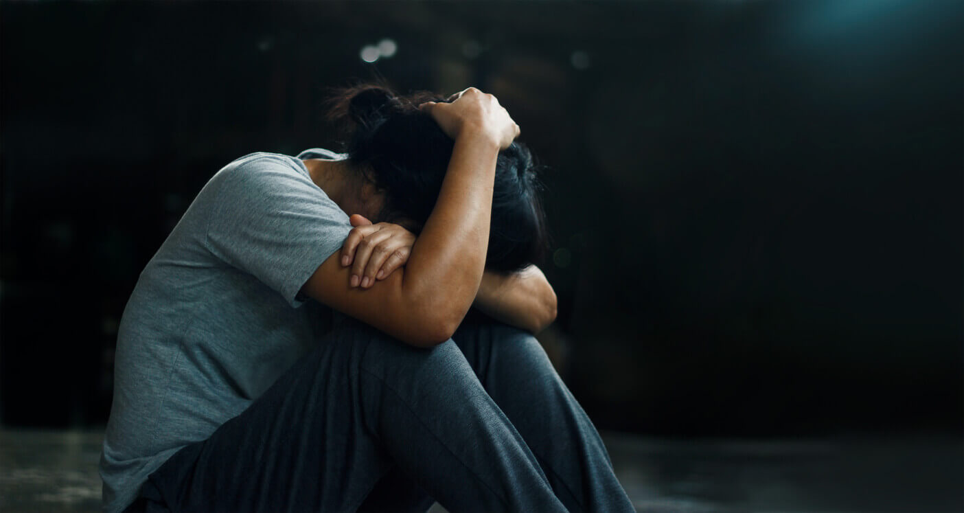 A woman with Major Depressive Disorder sits alone in a dark room, holding her head with her face down.