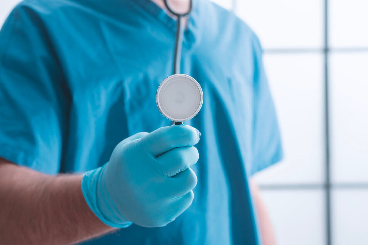 A close-up of a holistic primary care doctor in blue scrubs and protective gloves holding a stethoscope.