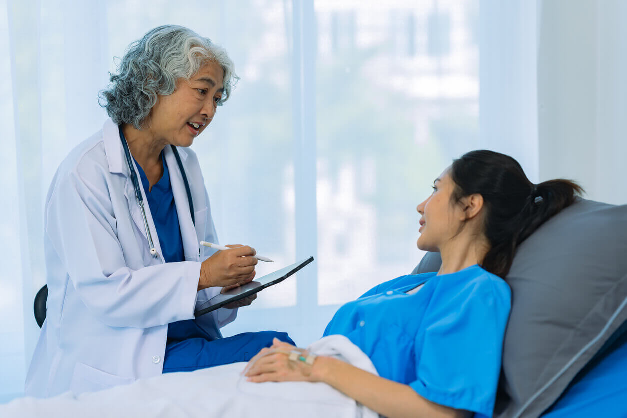 Elderly female holistic primary care doctor and Asian female patient are discussing something while sitting on hospital bed.